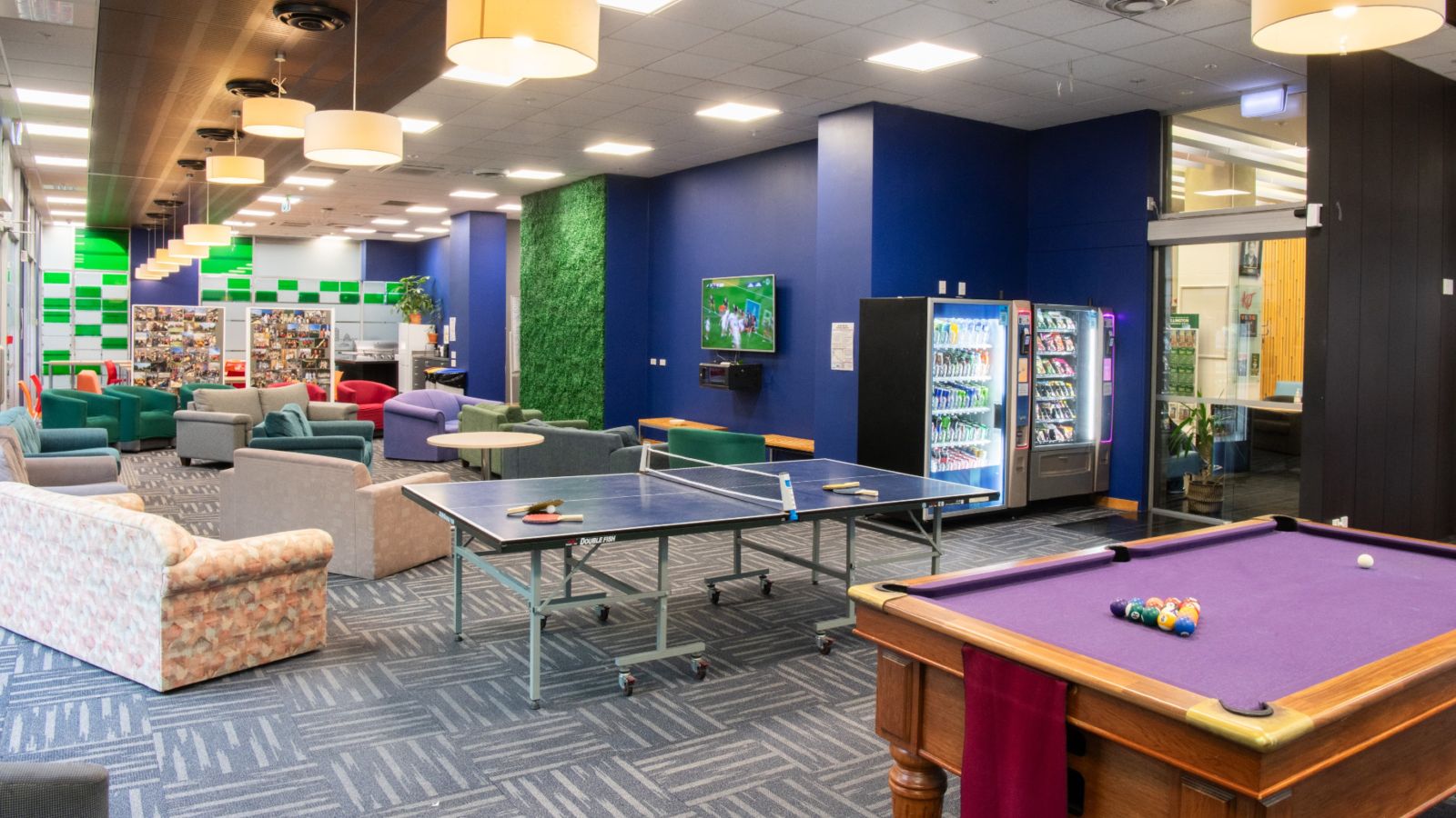 Large space shows pool table, table tennis, rows of sofas and chairs facing a large screen on the wall. Vending machines in the corner.