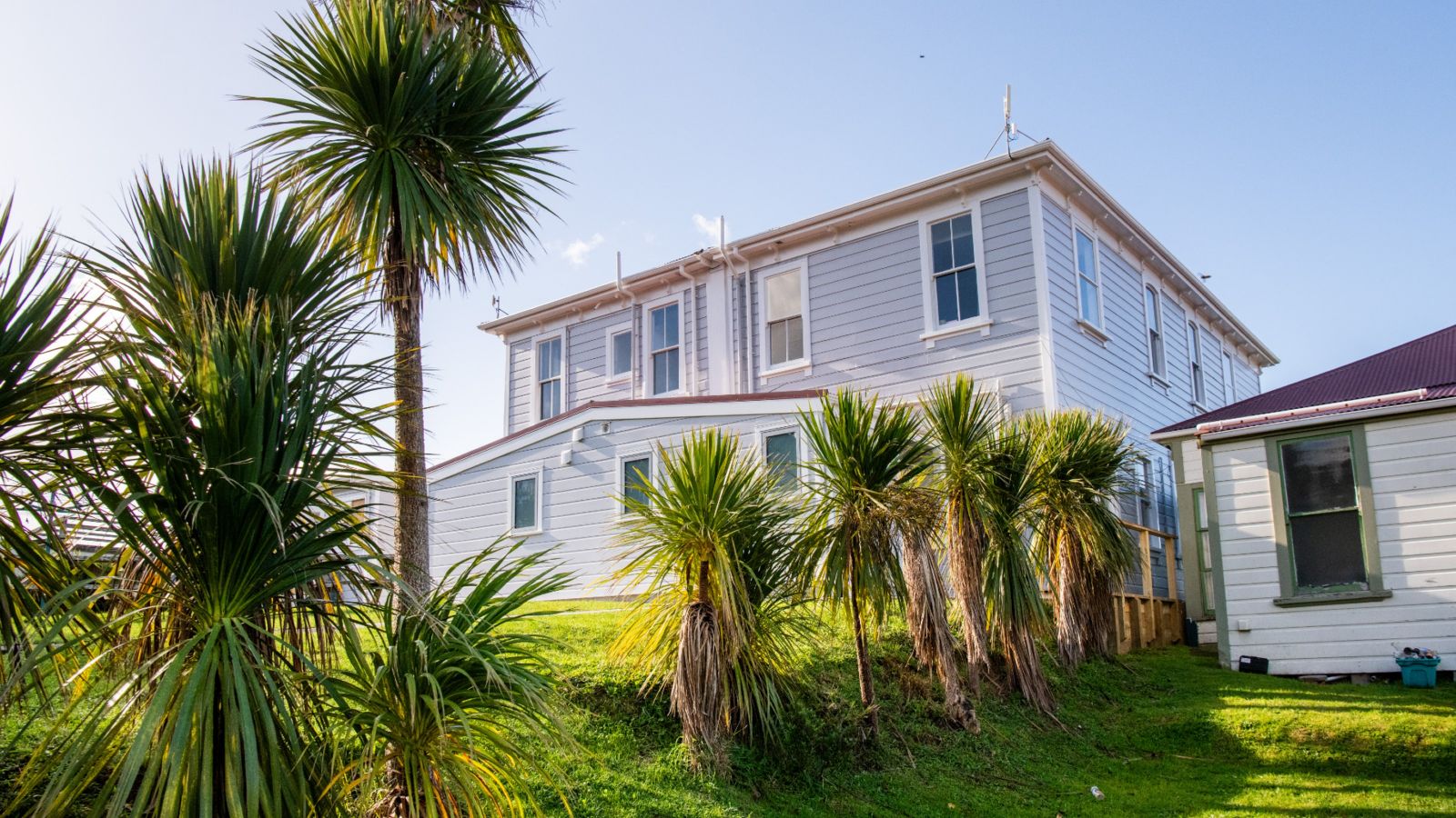 Large house with red roof, many windows stands in sun. Line of trees grow out of large grassy lawn.