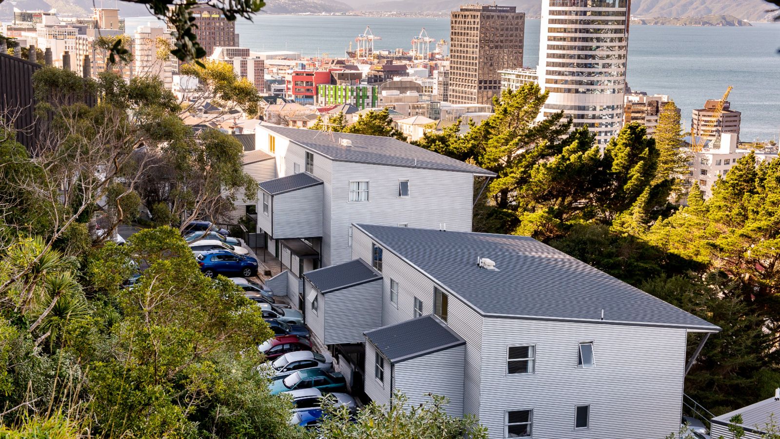 Two-story apartment buildings with cars parked in front, trees surrounding the buildings, and a view down the hill of the city