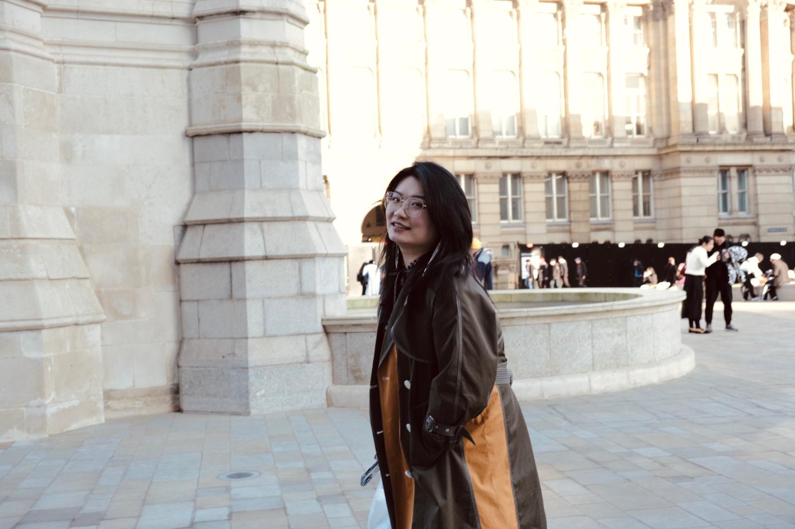 A woman with short black hair and glasses stands in a public square wearing a long two-tone trench coat&mdash;dark green on one side, orange on the other. Behind her rises a large stone building with columns and multiple windows; other people walk in the background.
