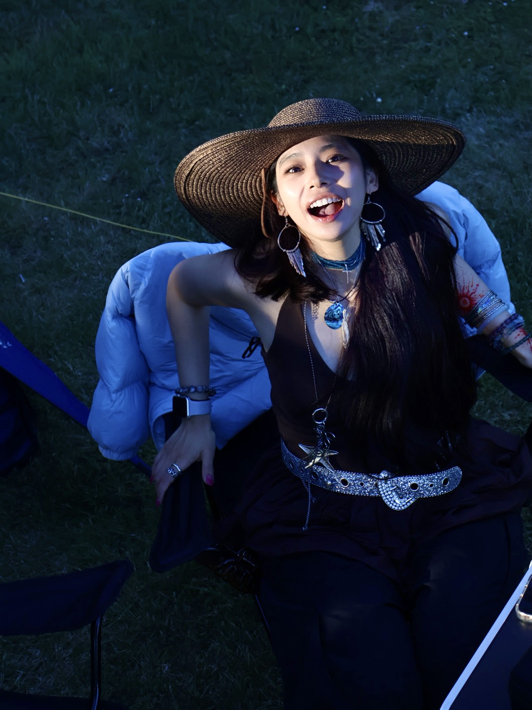 woman with tan skin and dark hair, wearing a wide-brimmed hat, visible jewelry (such as earrings or a necklace), and a dark-colored outfit. She is seated on a grassy outdoor surface, with a folded or draped jacket placed behind her. Her expression is playful and engaging, and she is looking upward&mdash;directly at the camera&mdash;with warmth and confidence. The setting appears to be a sunlit, natural environment.