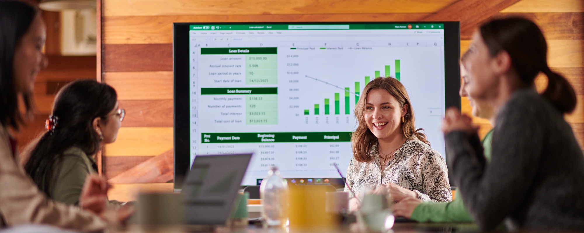 A group of people having a meeting around a wooden table with a large screen on the wall in the background.