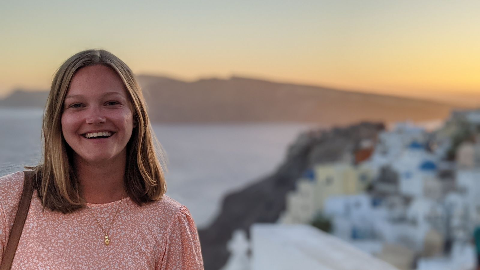 Head and shoulders photo of Olivia. Olivia has blonde hair and smiles at the camera and is wearing a pink shirt and a gold necklace. Out of focus behind Olivia is water and blue and white buildings.