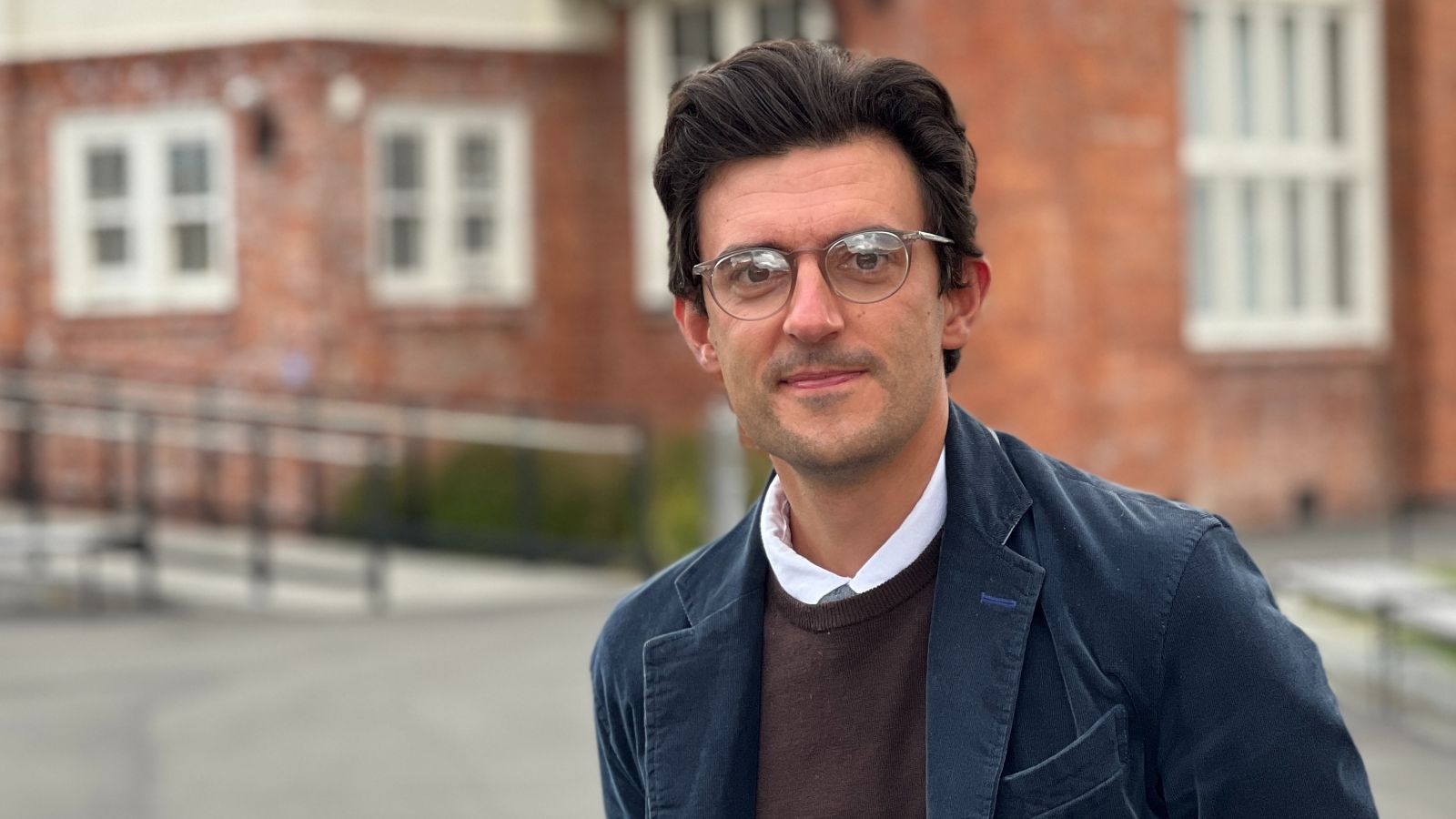 Costas has brown hair and wears glasses, a navy coat, and a dark sweater. In the background is a brick building.