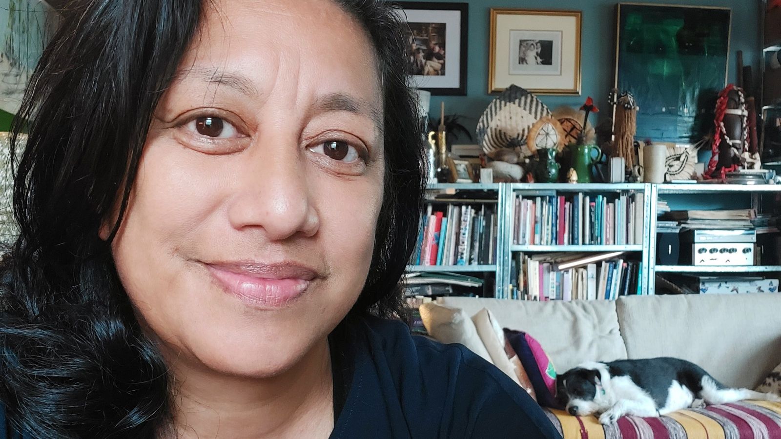 Head and shoulders portrait of Mishelle. Mishelle has dark hair and brown eyes and smiles at the camera. In the background a dog lies on a couch. Bookshelves and photos line the rear wall.