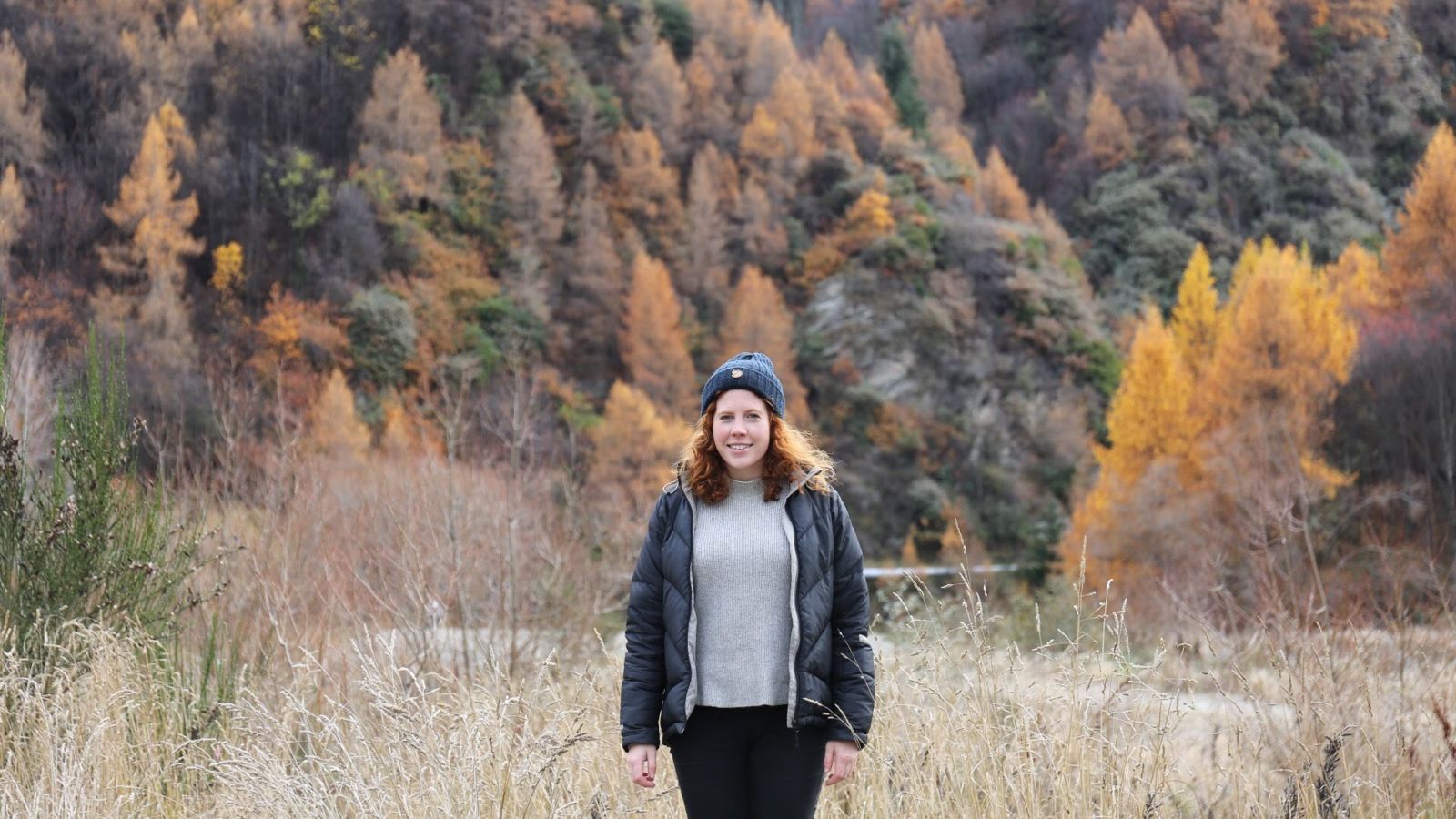 Sarah Rowley Adams stands in an autumnal forest, smiling at the camera. Sarah wears a grey sweater, black puffer coat and dark beanie. 