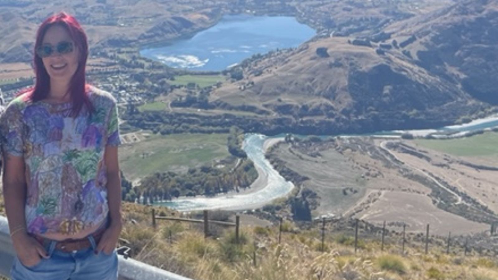 Photo of Tina Chiles in front of a pastoral scene. Tina has red hair and wears sunglasses, a multi-coloured shirt, and jeans.