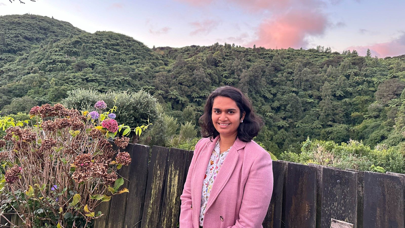 Photo of Ushana Jayasuriya. Ushana wears a pink blazer and smiles at the camera. Ushana stands in front of a fence. In the background is a forested hill.