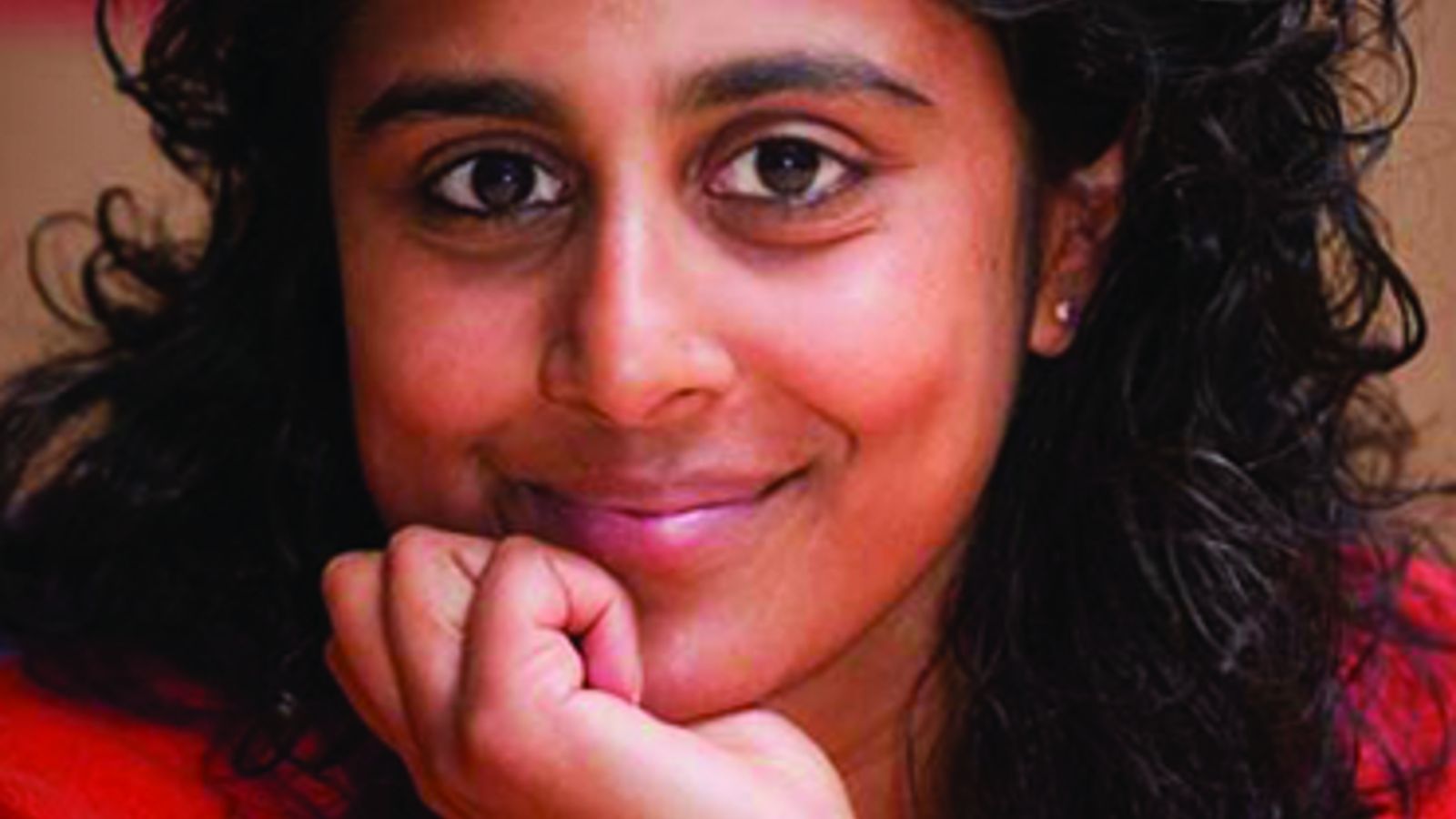 Photo of Anushka Brooking née Perinpanayagam. Anushka has dark, curly hair and holds her chin in her hand.