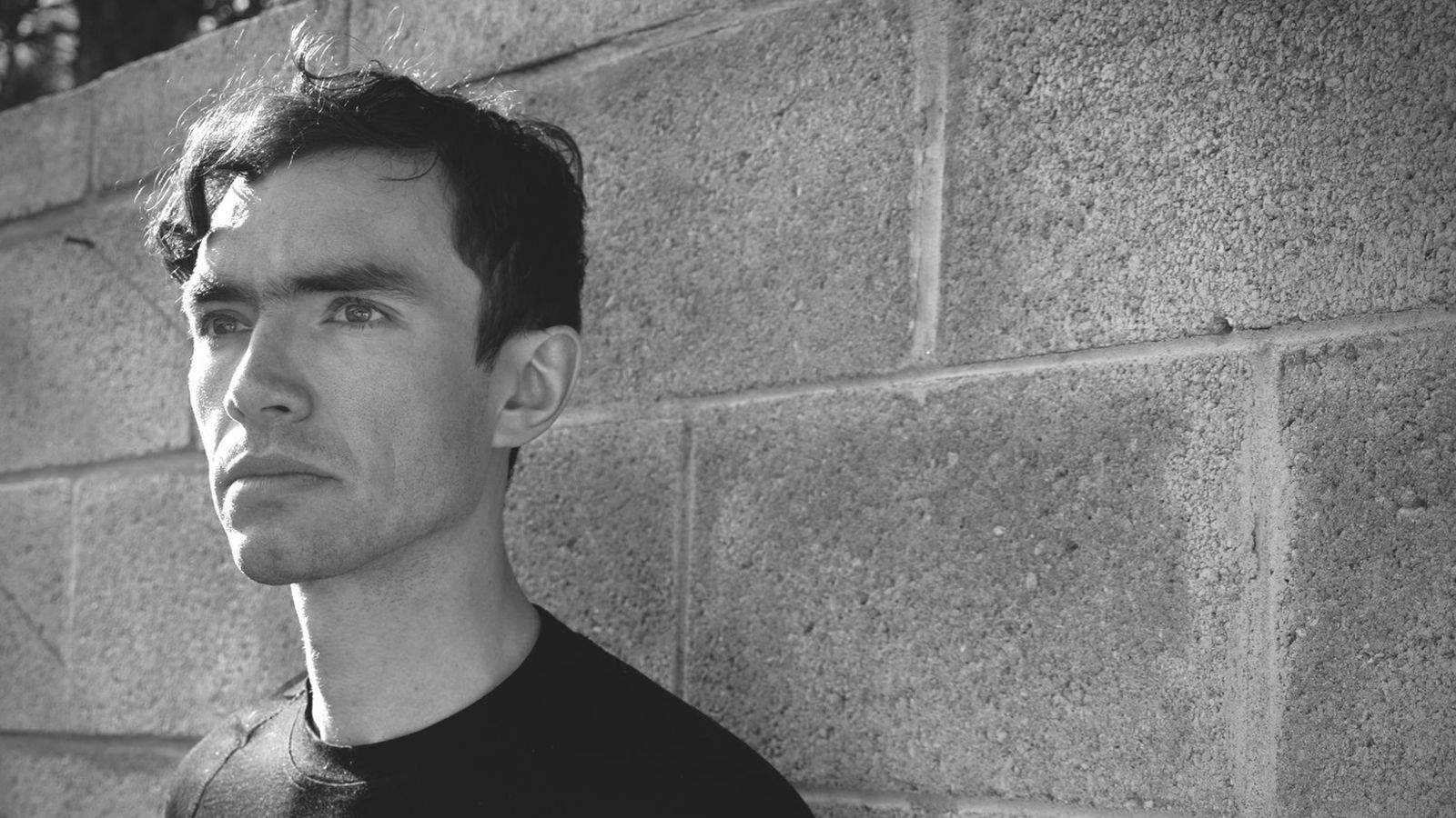 Black and white head and shoulders portrait of Jim Murphy. Jim stands in front of a concrete wall and looks off camera.