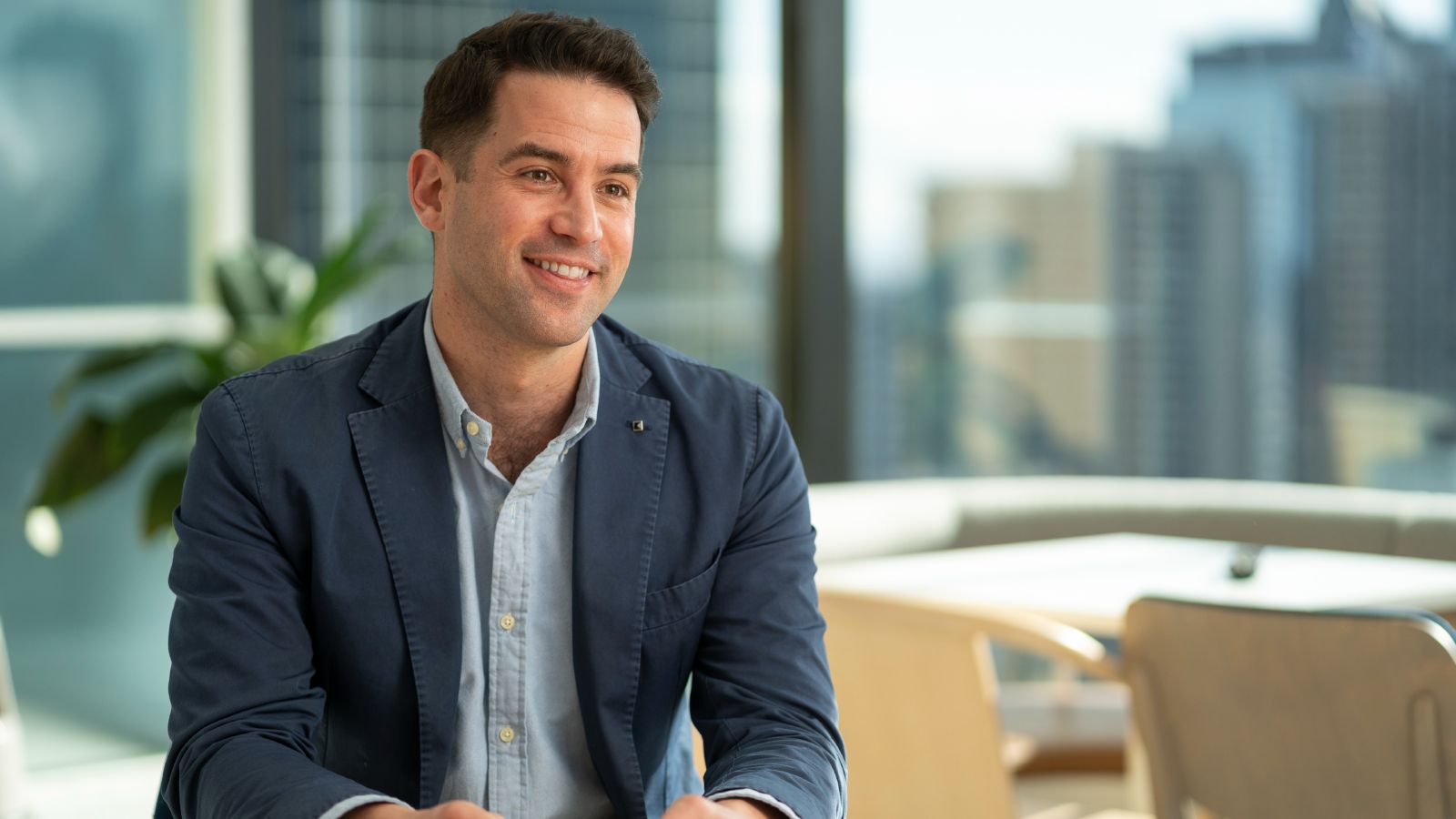 Portrait of David Herman, who smiles and looks off camera. David has brown hair and wears a dark blue jacket over a light blue shirt. A skyline of tall buildings can be seen in the out-of-focus background.