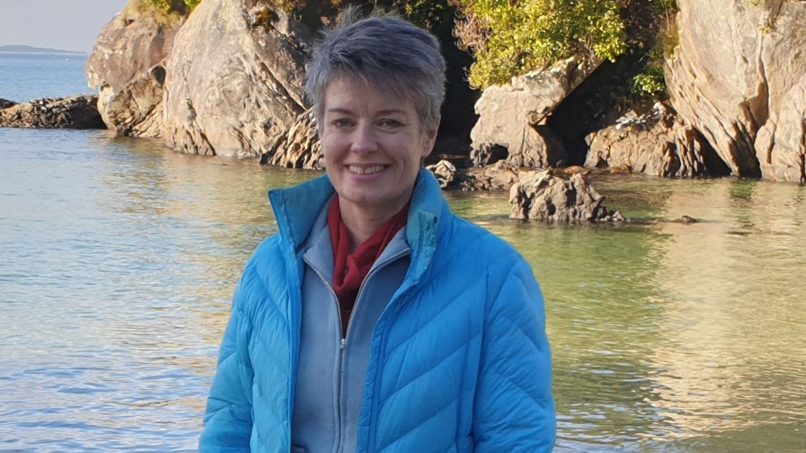 Photo of Catherine Duthie in a blue jacket in front of water and a rockface.