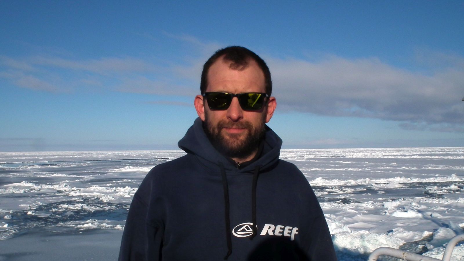 Head and shoulders portrait of Flavio Mayorga. Flavio wears a navy blue hoodie and sunglasses, sea ice in the background.