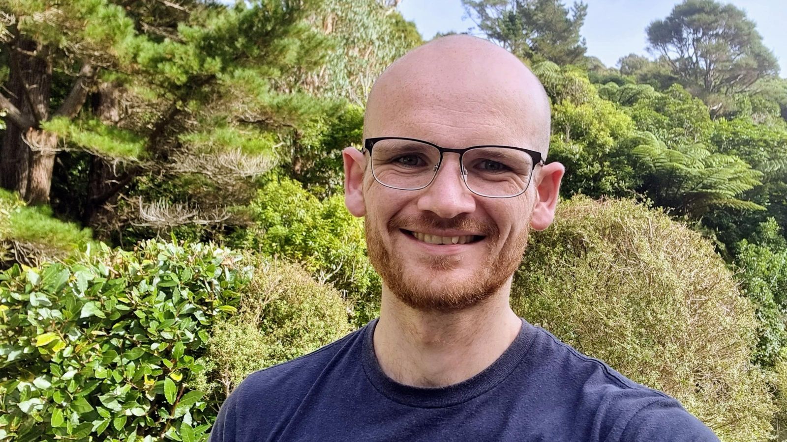 Head and shoulders portrait of Thomas Butt smiling at the camera. Thomas wears glasses and a blue shirt and stands in front of greenery.