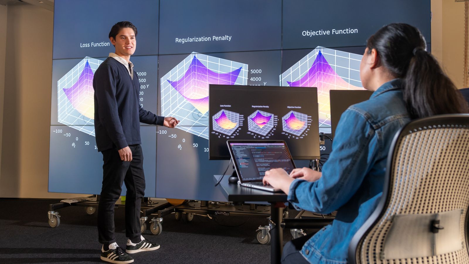 Student stands in front of multi-screen display while a second student operates a laptop