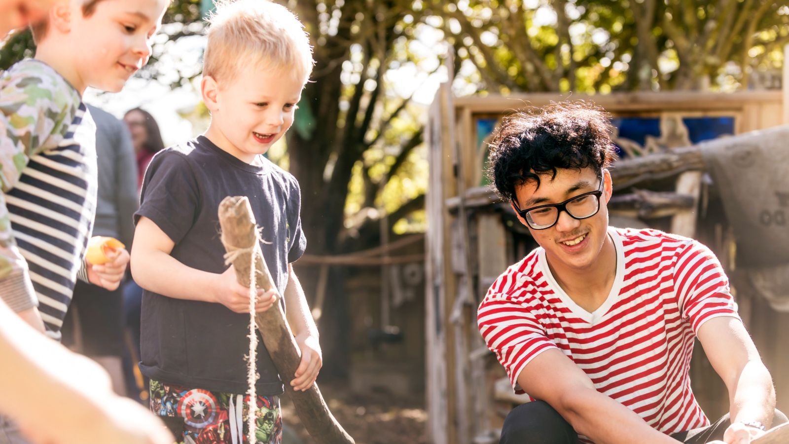 Two preschool aged boys holding sticks watching a male ECE teacher do something