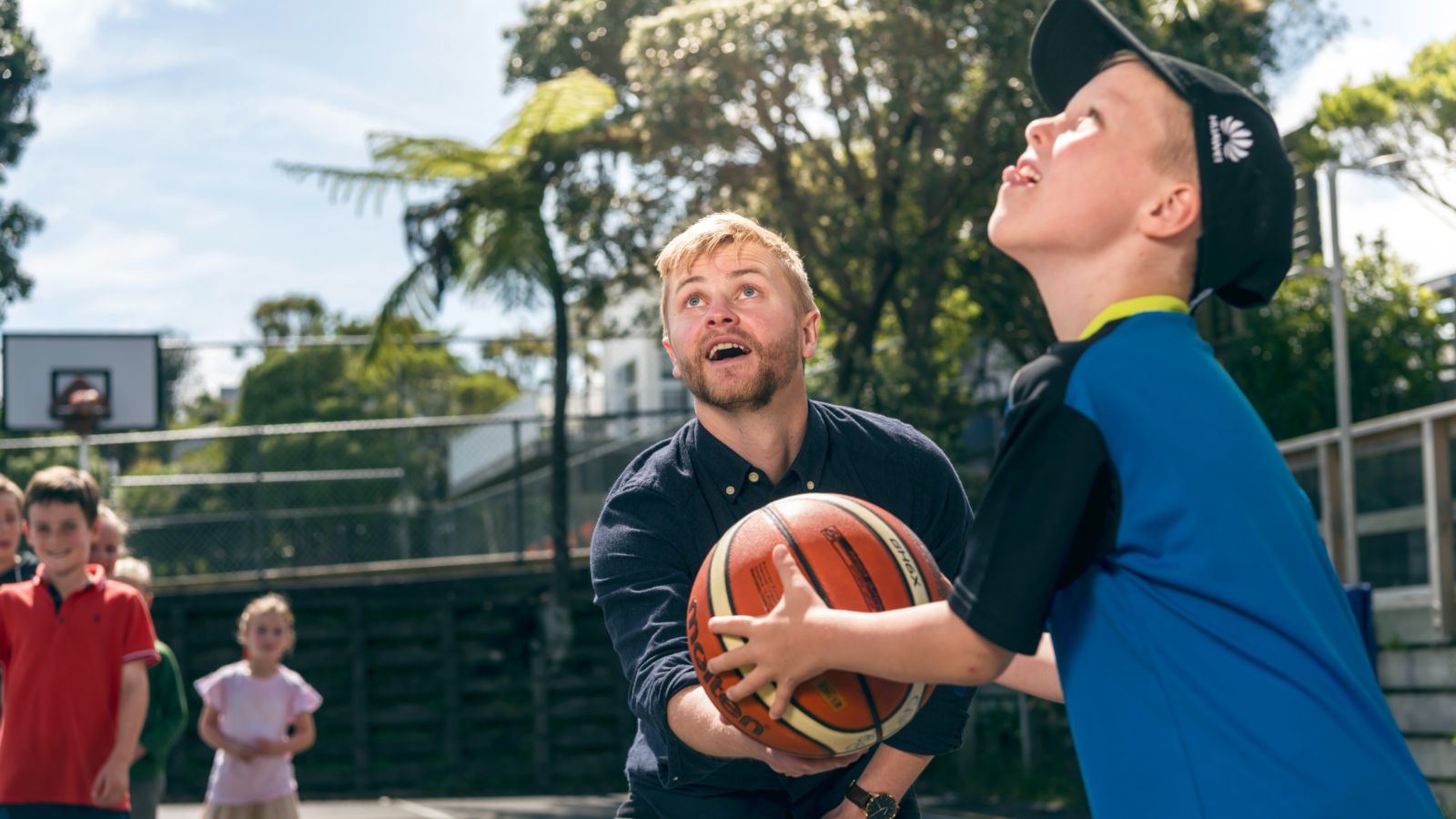 Male primary teacher teaching a boy in a blue shirt how to shoot a basketball