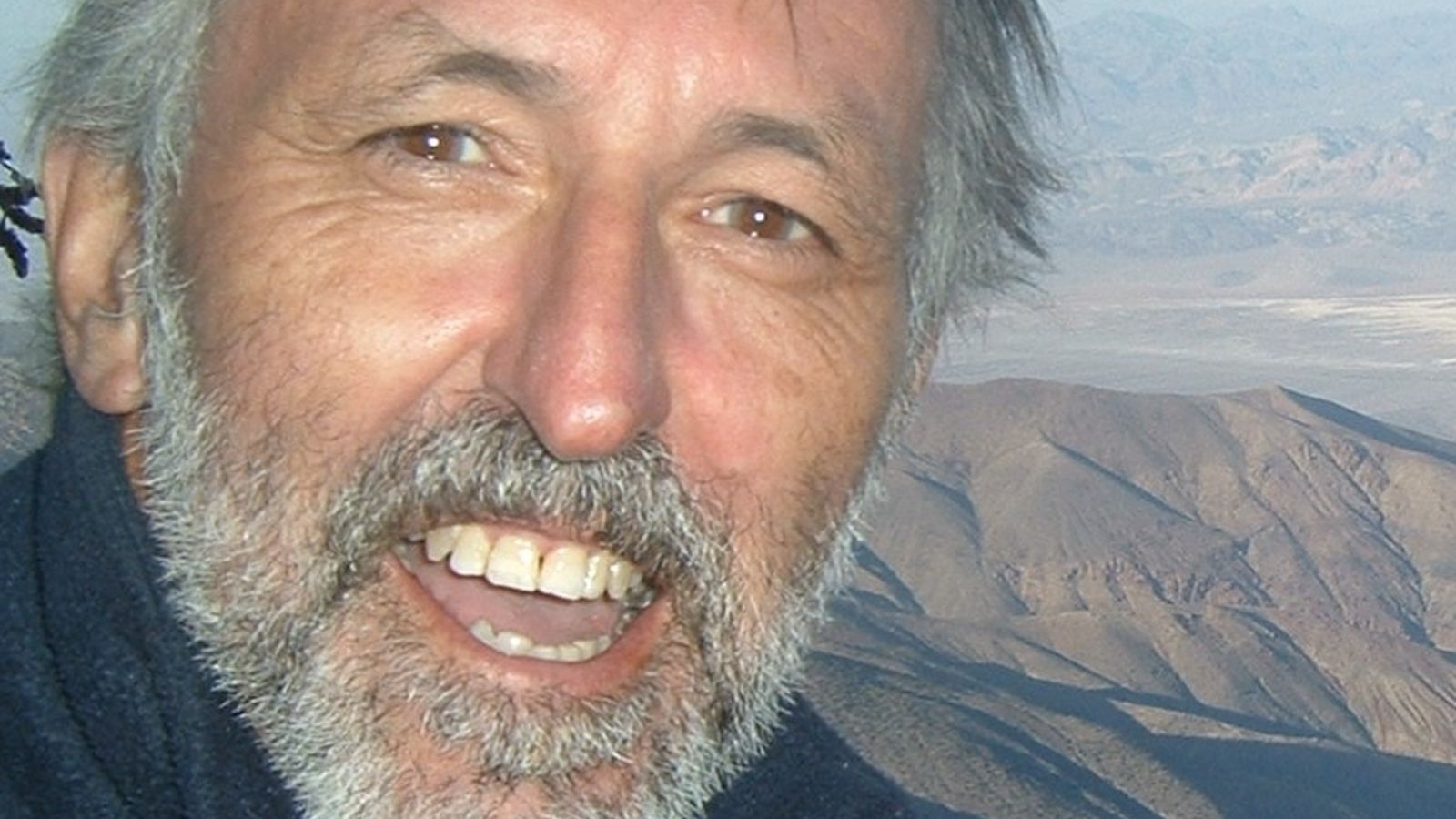 A man with grey hair and a beard looking at the camera. There is a view of mountains behind him.