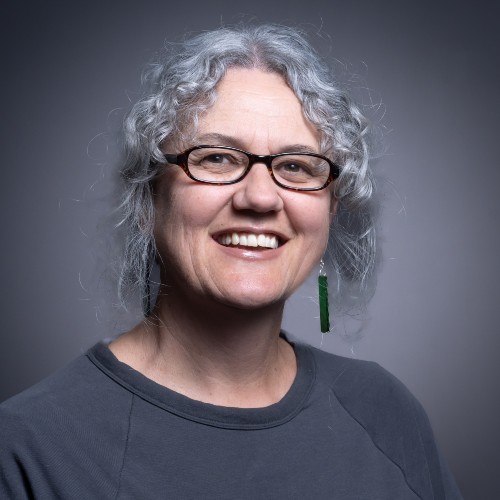 Māori wāhine (woman) with small glasses and grey curly hair smiling. 