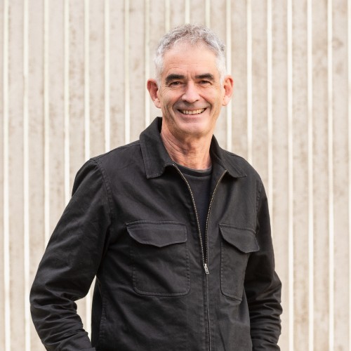 Man with grey/white hair wearing a black shirt with hands in his pockets smiling with teeth.