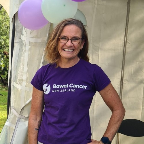 Woman with short blonde hair wearing black rimmed glasses and a purple t-shirt smiling.