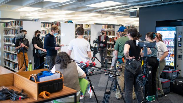 A film crew setting up cameras and equipment for filming in a library next to shelves full of books.