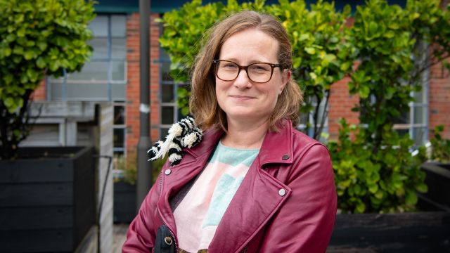 A woman stands in a garden. She is wearing glasses and a red jacket and is looking directly at the camera.