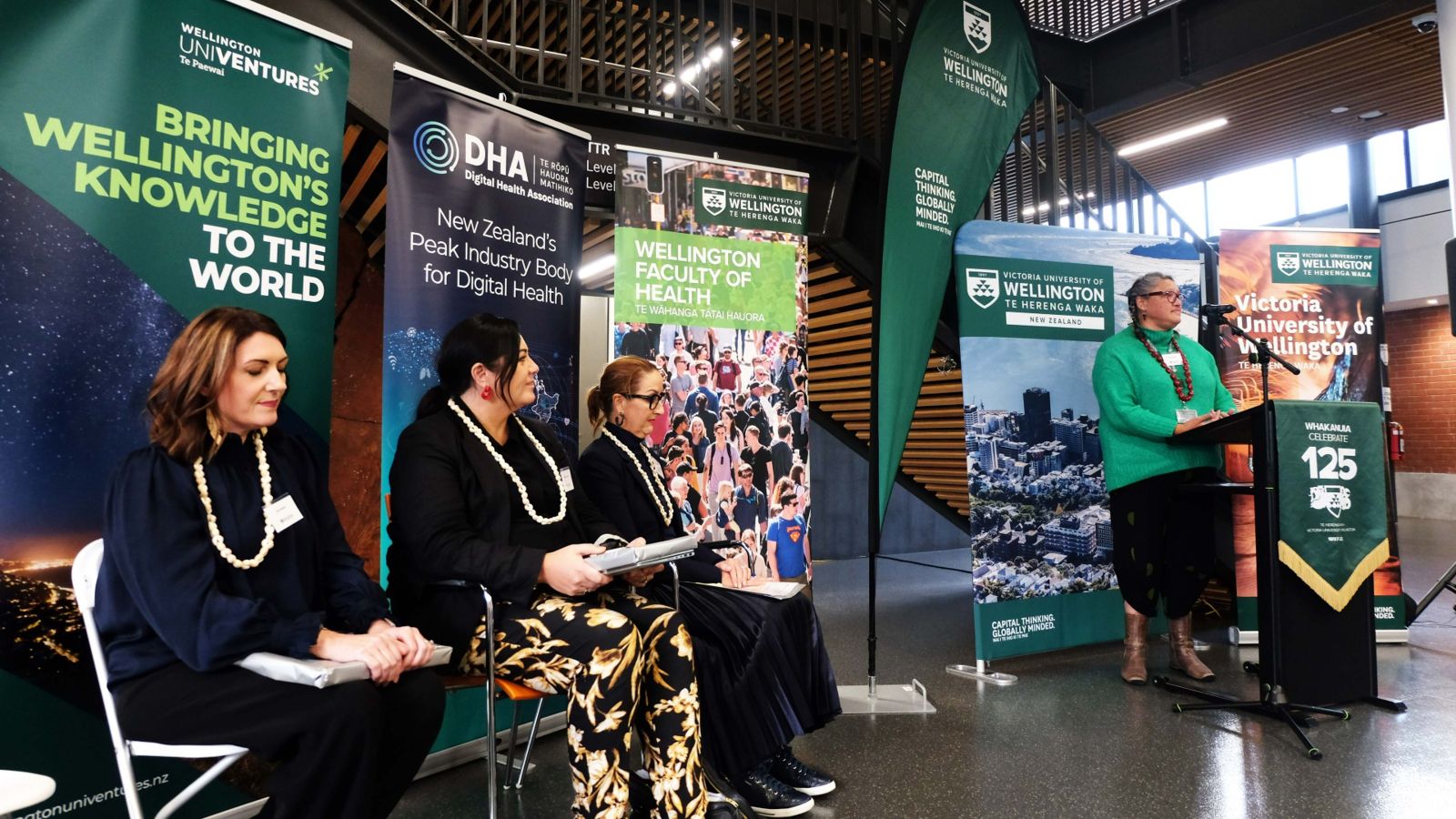 Fran Kewene stands at a speakers lectern delivering a speech to a crowd that is not visible in the image. The three speakers from the event sit alongside her listening intently.