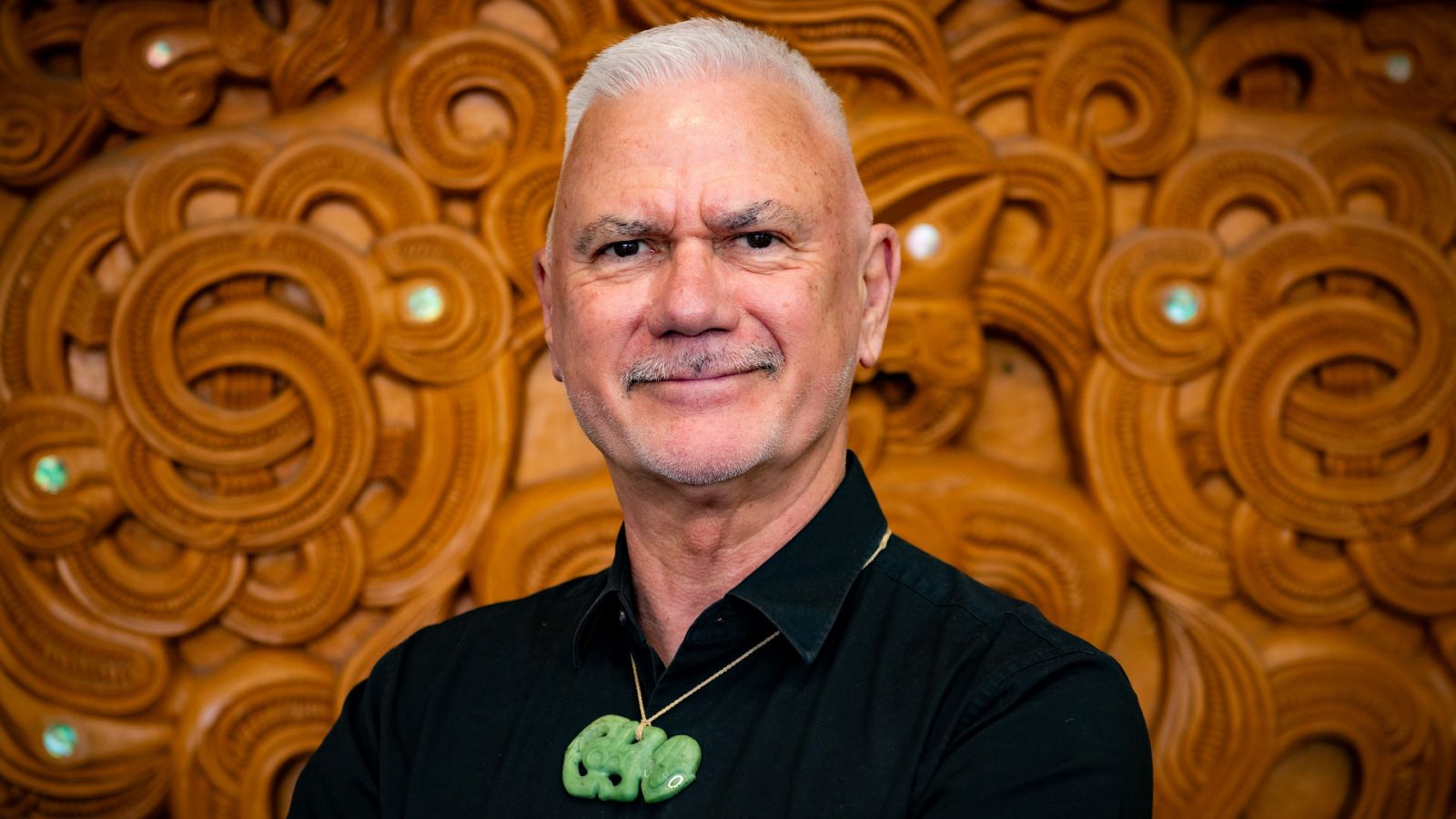 Man with Pounamu greenstone pendant stands against wooden Maori artwork backdrop, showcasing cultural adornment and traditional art.