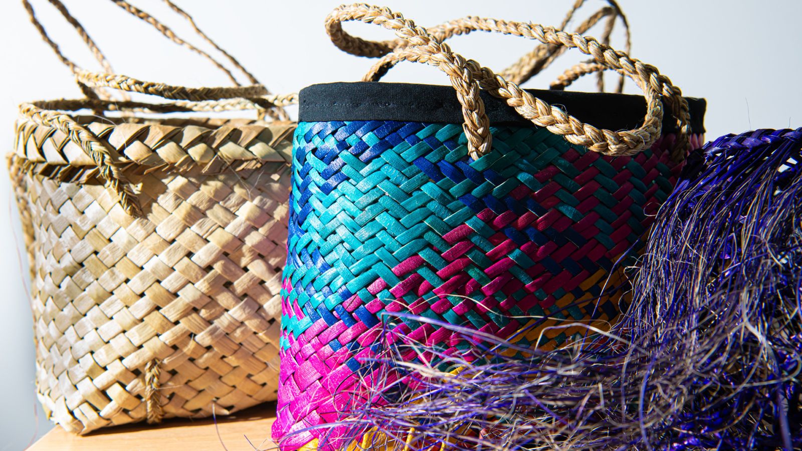 Two kete or woven baskets. One is purple, pink and blue and the other is traditional brown.
