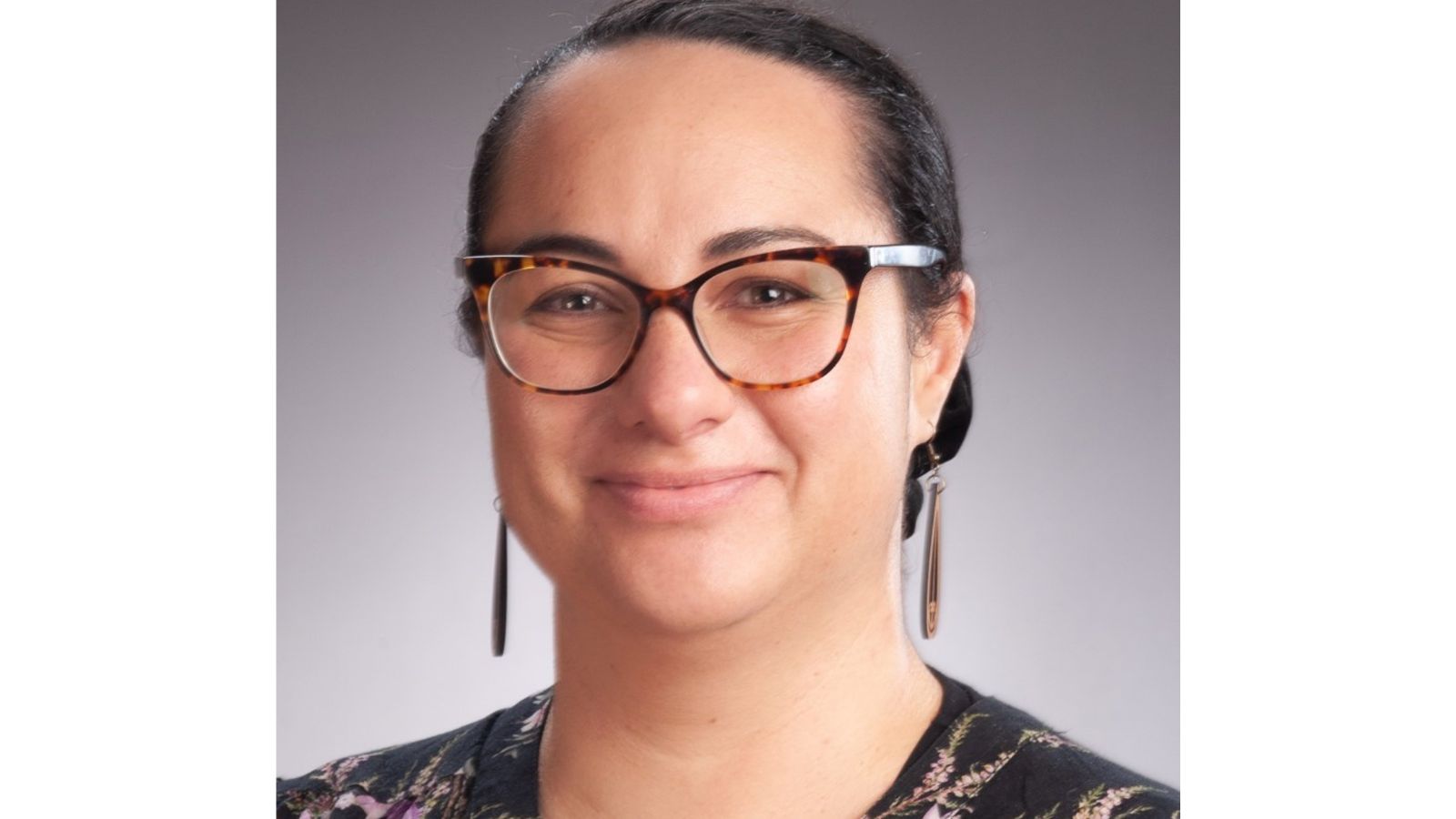 A portrait of Ngaire. She is wearing her hair up and has long earrings on. She is wearing black framed glasses and is smiling.