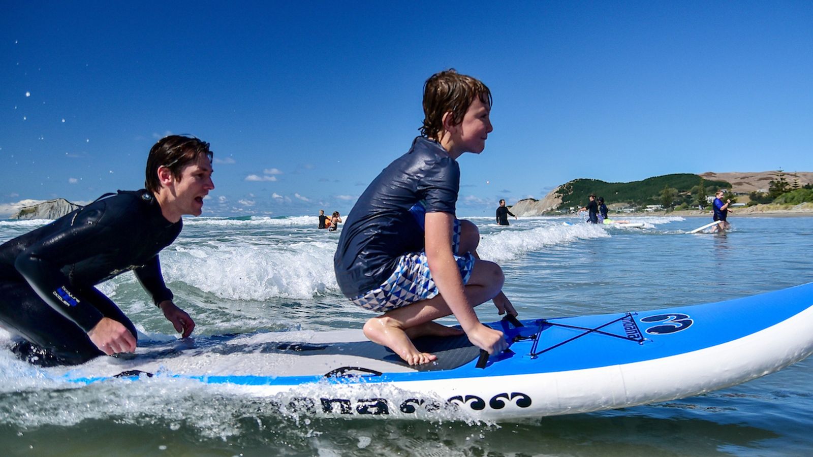 Two young boys on a paddleboard in the surf. They are mid action catching a wave. The younger one is wearing a rash vest and board shorts and the older one is wearing a wetsuit. 