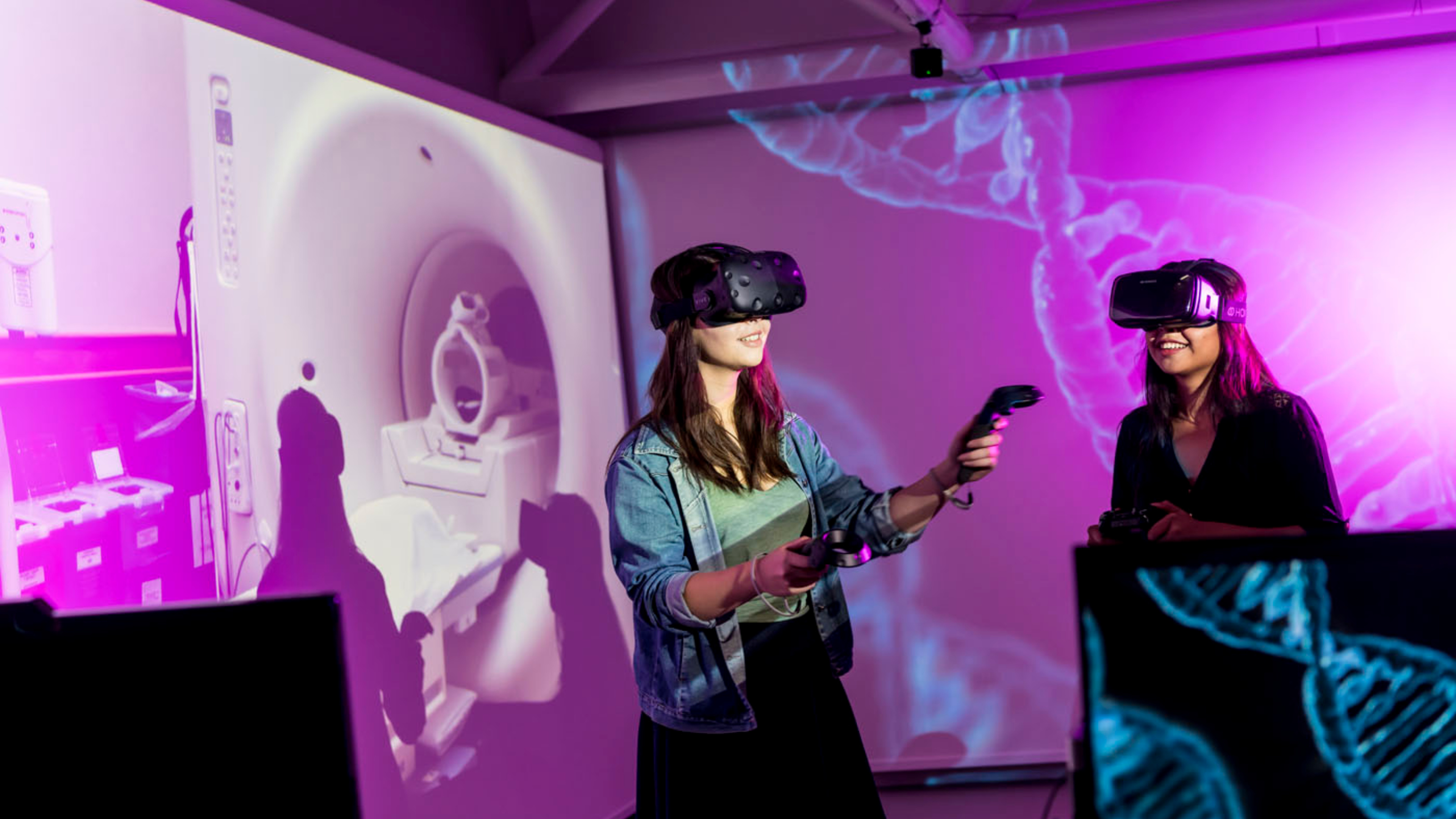 Two women using Virtual Reality headsets in front of a projected image of DNA helices and an MRI machine