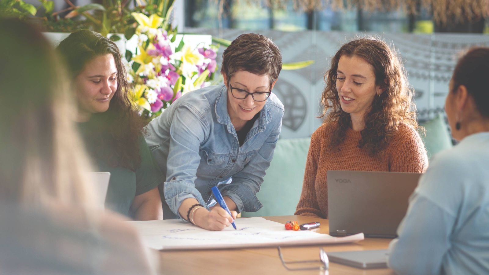 Four people collaborate around a table with paper, laptops, and stationery in a bright, plant-filled room—likely a health or wellbeing setting.