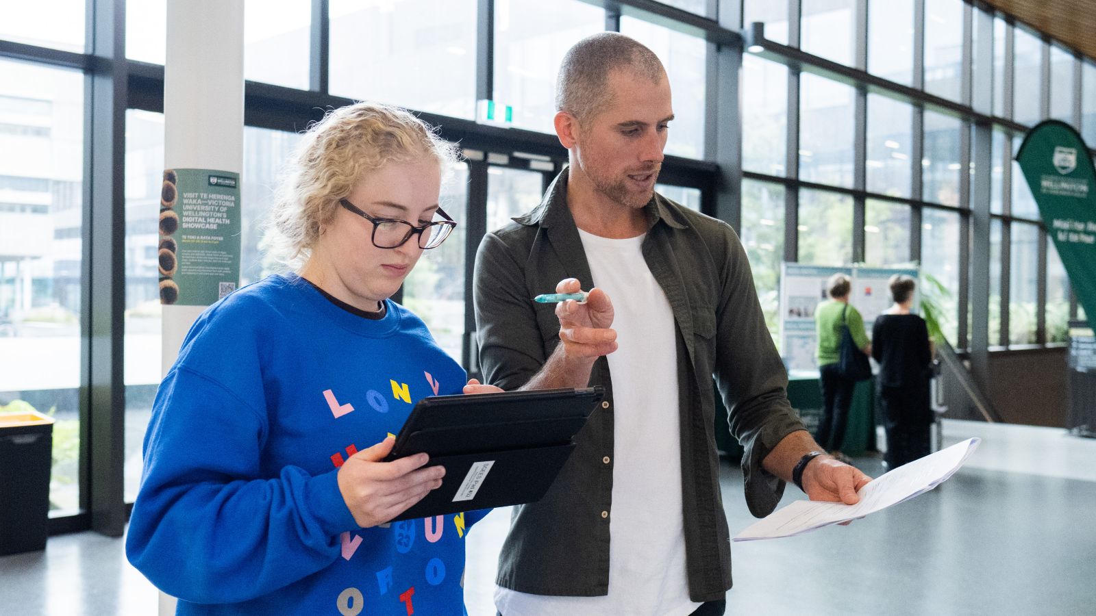 A man stands next to young woman. The young woman is holding a tablet device and looking intently at it. The man is gesturing with his hands while holding a pen and reading a piece of paper. It looks like he is explaining how something works to her.