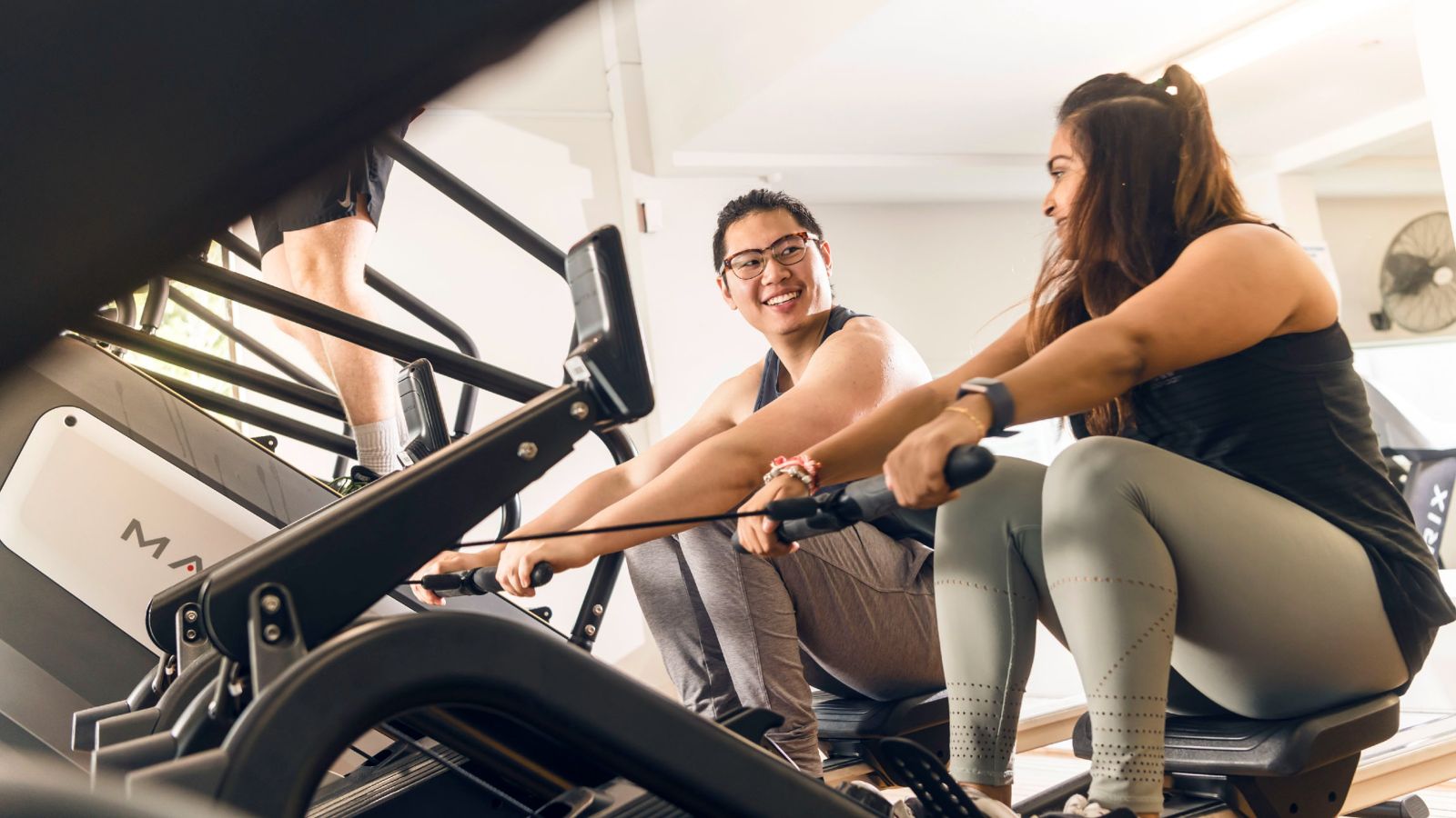 Two people on rowing machines in a gym, they are looking at each other as if having a conversation while exercising