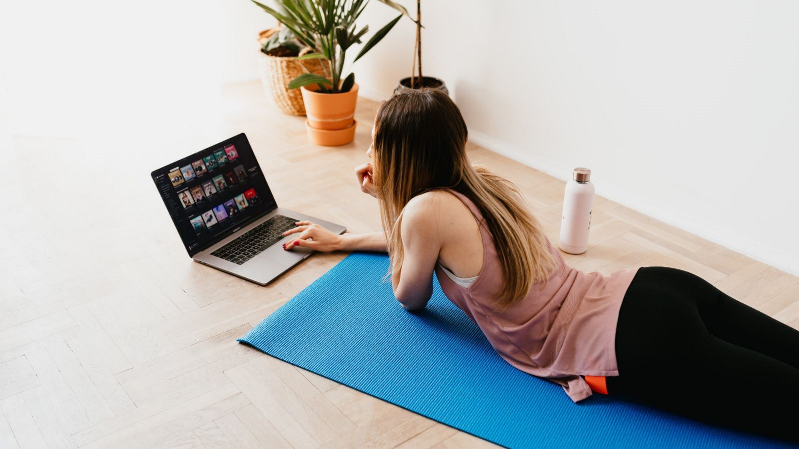 A woman lying on a yoga mat in active wear, looking at a laptop