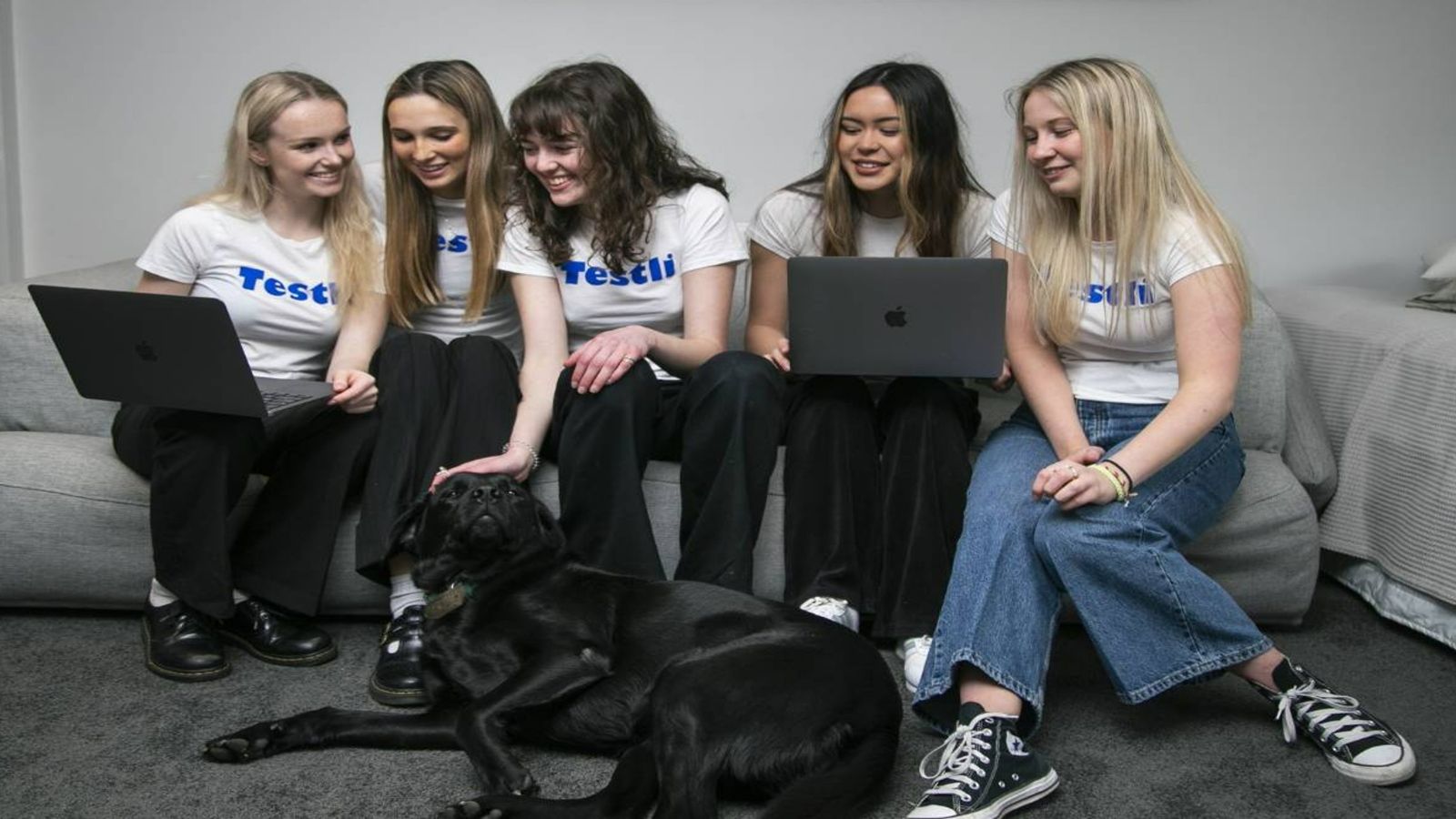 Five young women sitting on a couch working, with a dog
