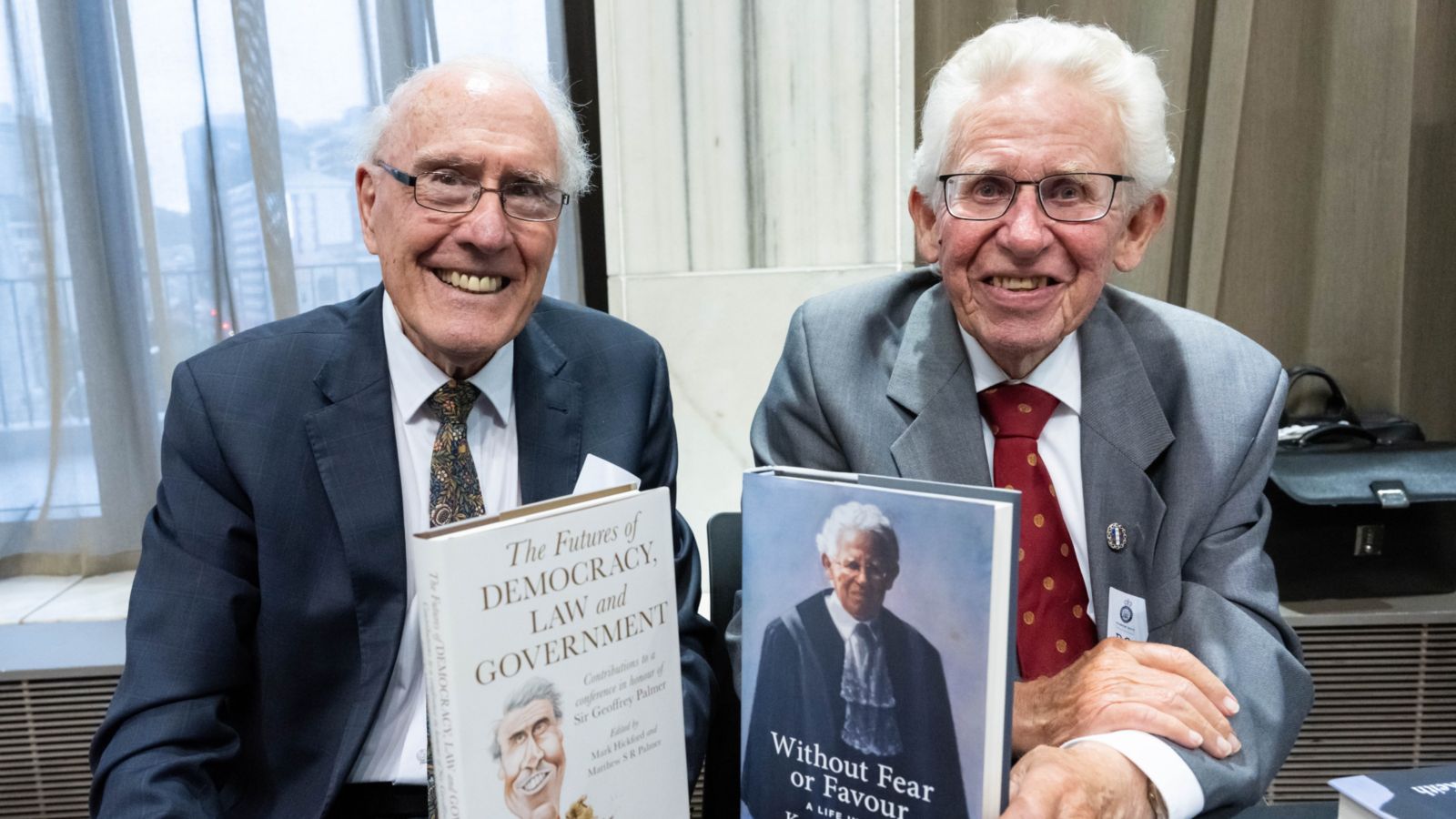 Two older men smiling at the camera, each holding a book.
