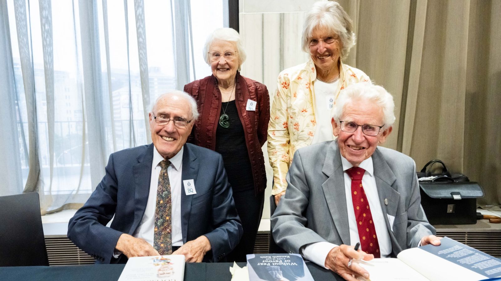 Two older men sitting at a table, with their wives standing behind them proudly.