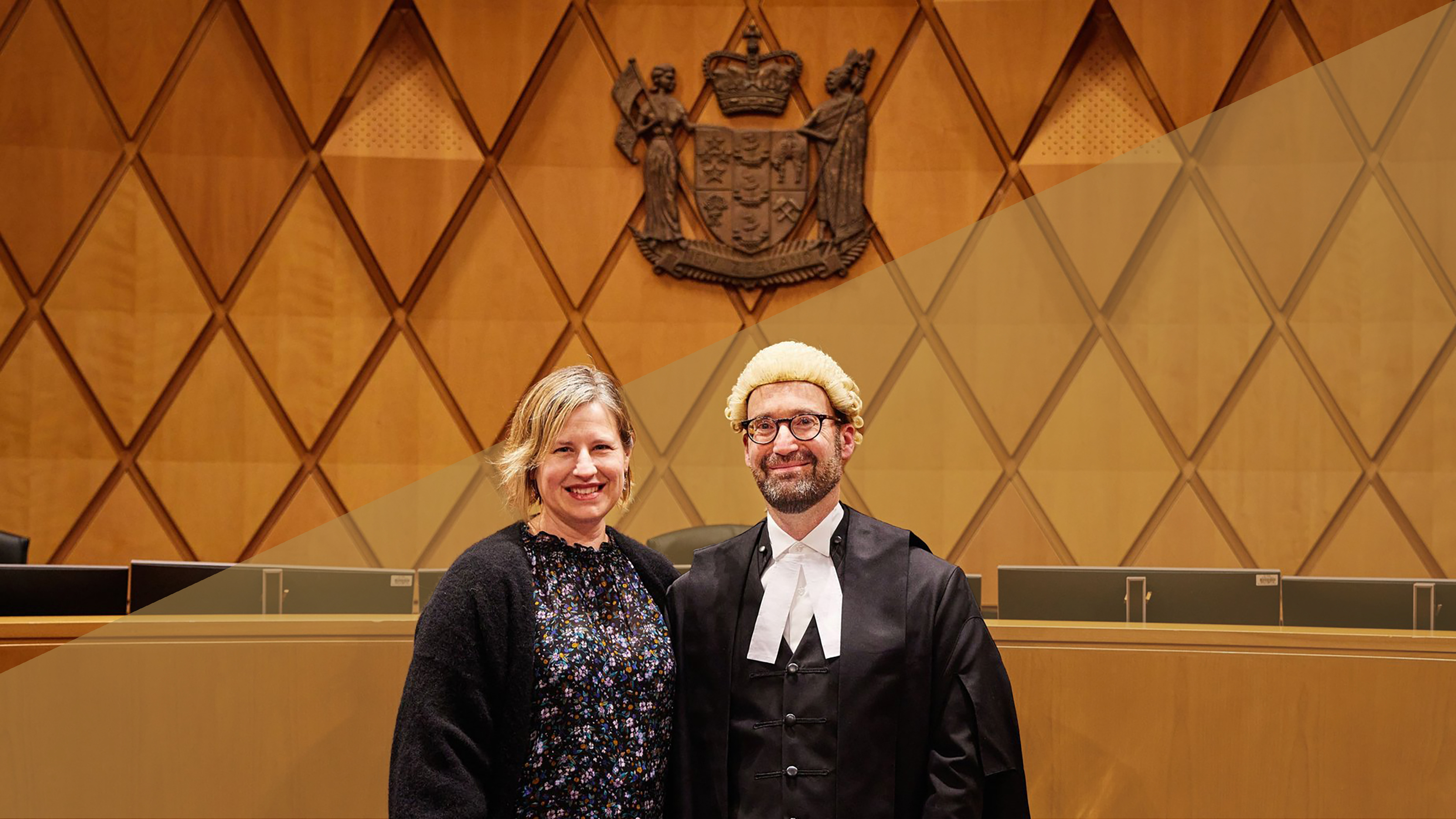 A man and woman in front of a wooden wall, he is wearing ceremonial legal robes.