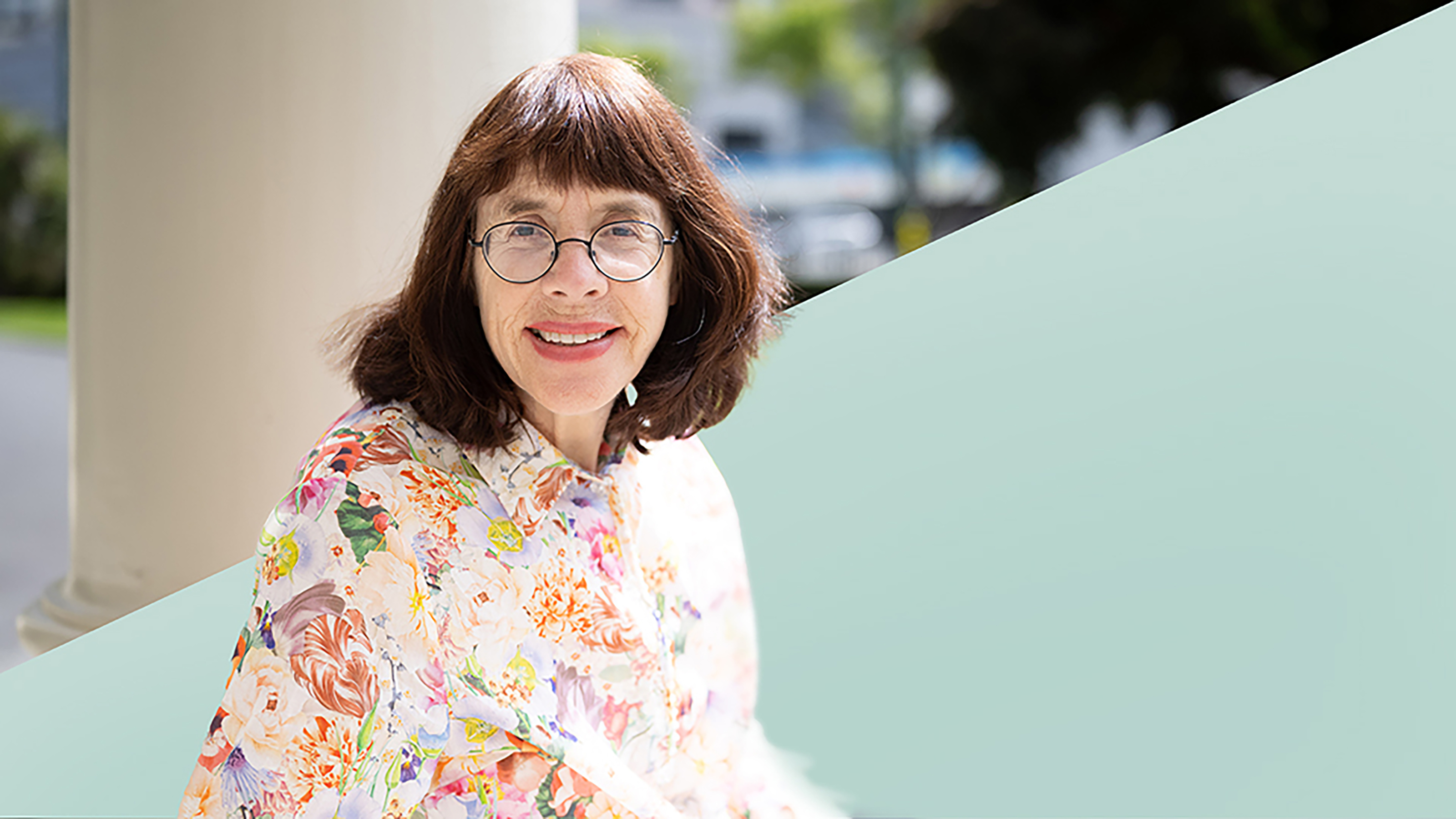 A woman in a floral shirt and glasses smiles to the camera. 