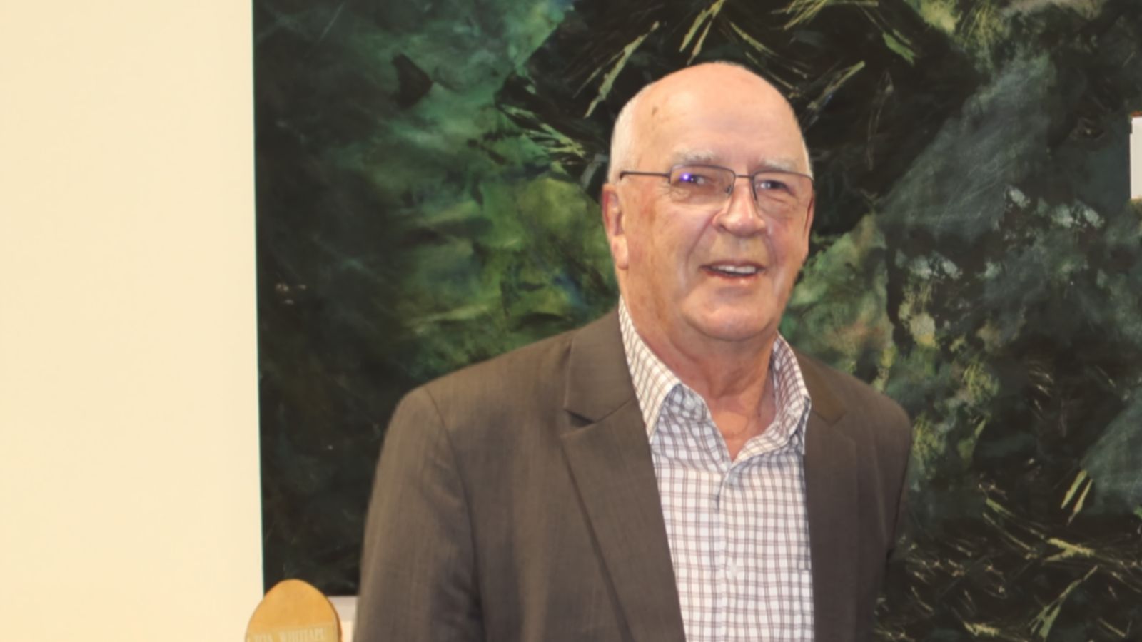 Man with grey hair stands with brown blazer looking at the Camera