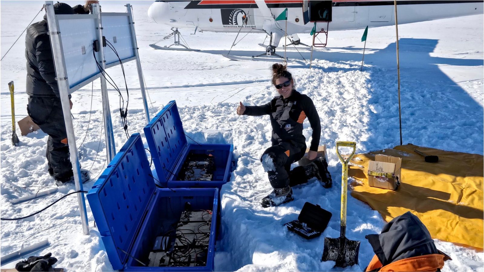 Girl doing thumbs-up on ice with science equipment around her and a plane in the background, in antarctica