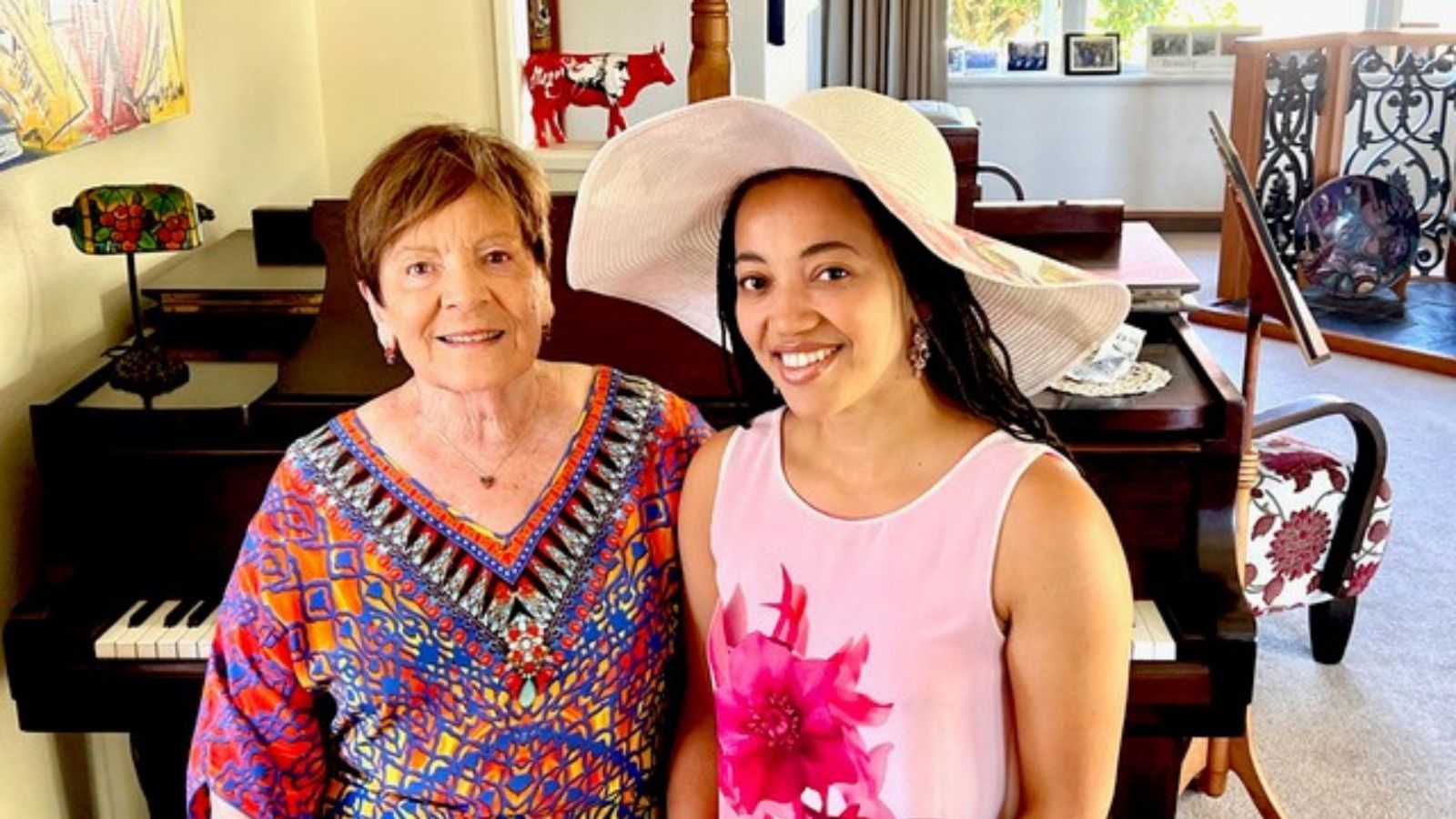 Two women, one older with short hair and a bright pink floral top, the other dark-skinned wearing a large hat, with black hair, wearing a pale pink singlet with a dark pink flower on it