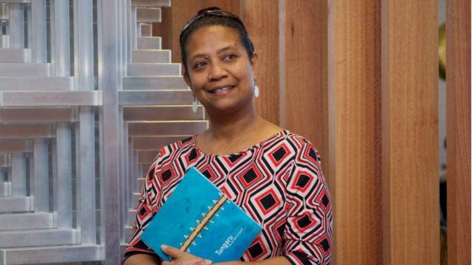 woman with dark hair pulled back, wearing colourful top, against background of wood and steel