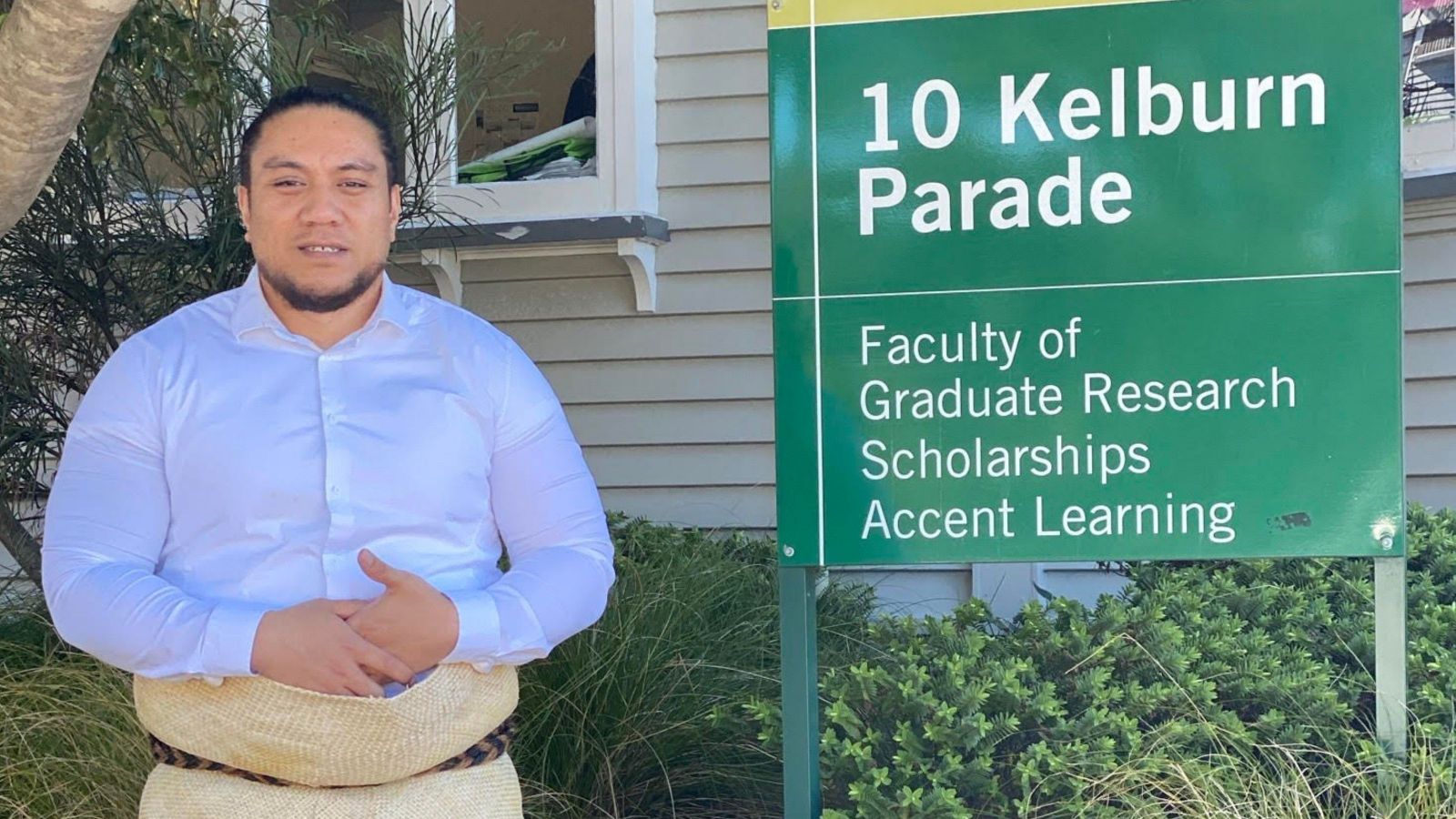 Man with tongan woven tupengu standing beside green sign which says 10 Kelburn Parade, Faculty of Graduate Research, Scholarships, Accent Learning