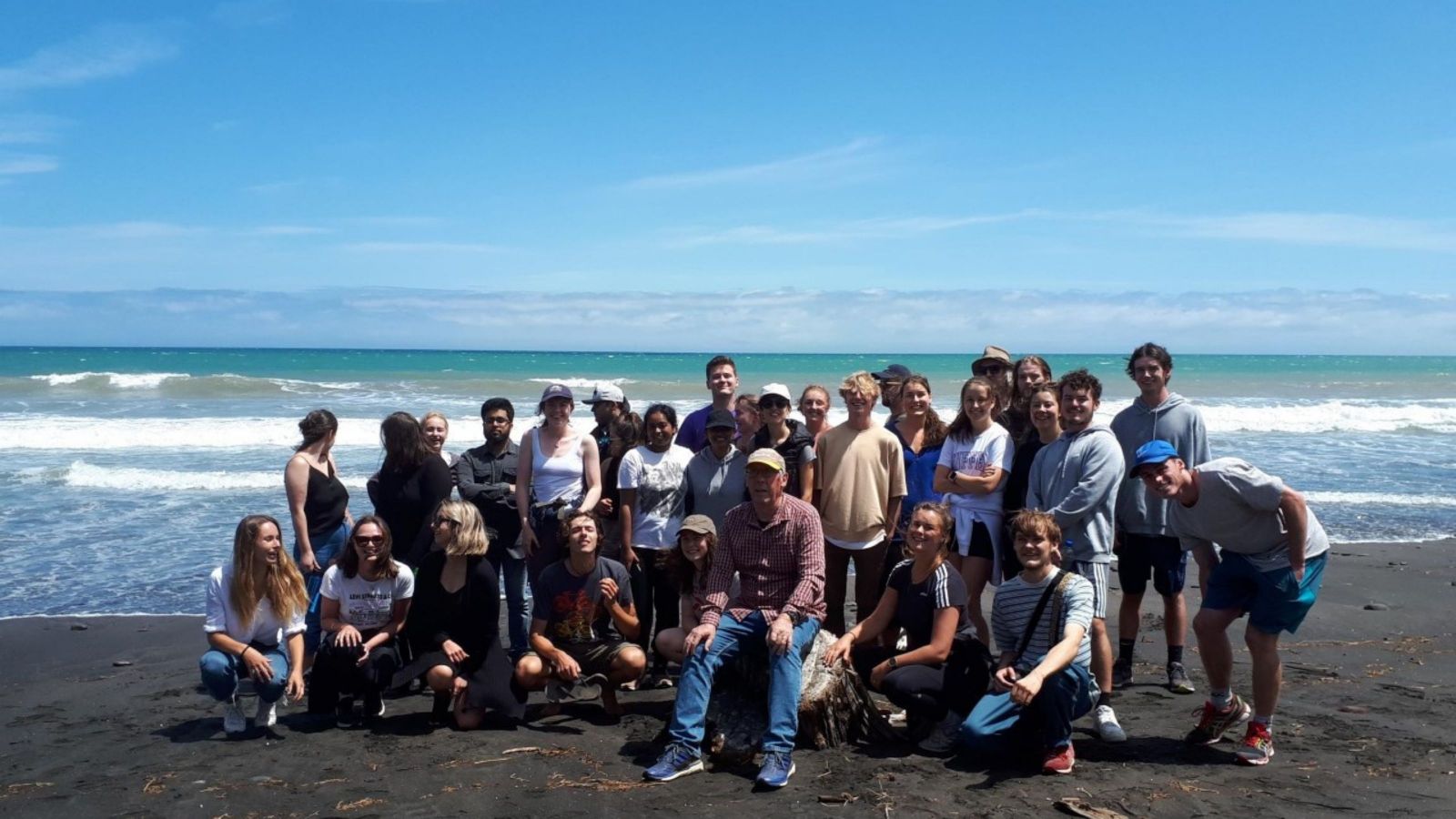 A group of around 30 people on a beach with blue sky and crashing waves behind them