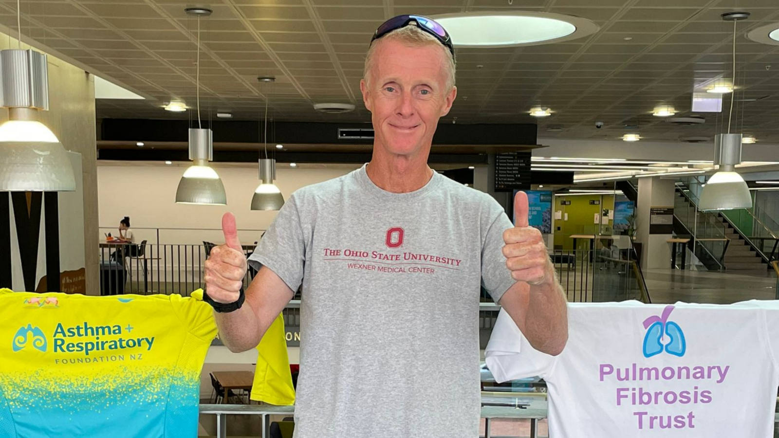 Marketing Professer Nick Ashill stands with two thumbs up in front of tshirts promoting the Pulmonary Fibrosis Trust and New Zealand Asthma and Respiratory Foundation