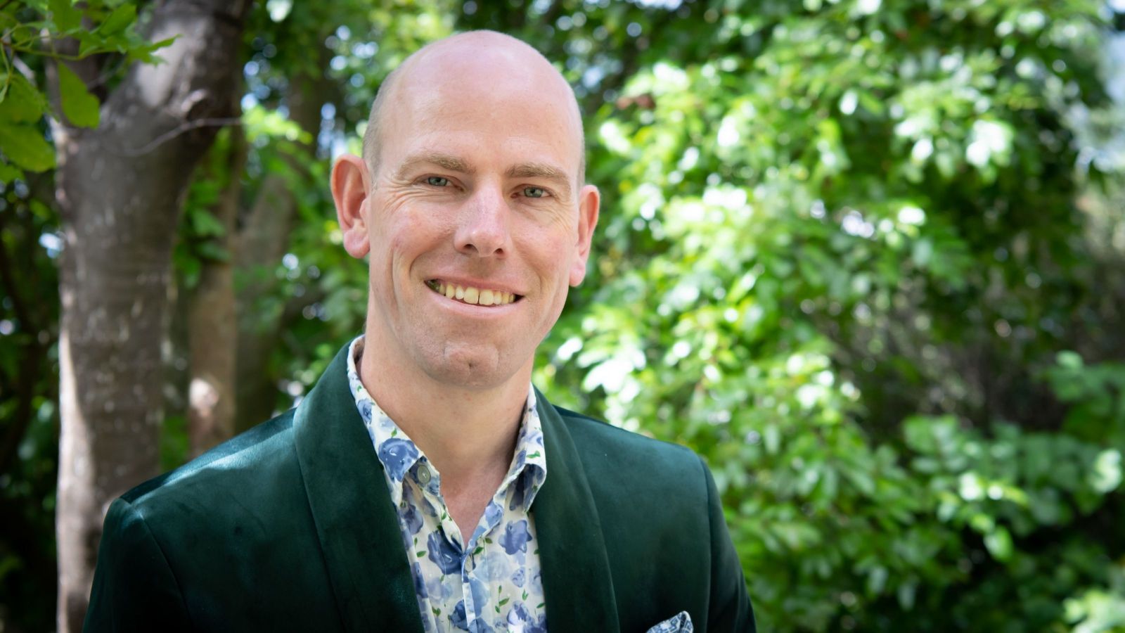 bald man smiling at camera wearing nice suit with trees in background